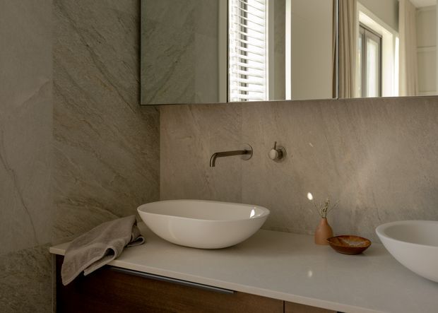 Close-up of a refined contemporary bathroom vanity with two white vessel basins on a pale stone benchtop, wall-mounted brushed metal tapware, and a large mirror. Full-height stone-look wall surfaces, warm timber cabinetry, and louvred windows create a calm, softly lit interior.