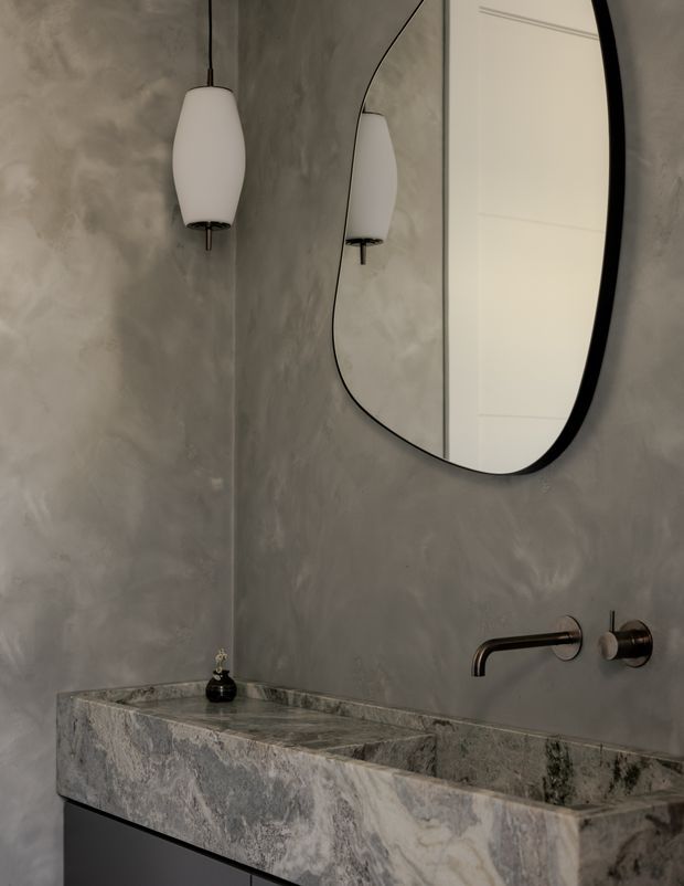 A moody, minimalist bathroom vanity featuring a thick natural-stone basin and benchtop with an integrated rectangular trough, wall-mounted brushed metal tapware, a large organically shaped black-framed mirror, and a sculptural pendant light. The walls have a mottled grey polished-plaster or concrete-like finish, with dark cabinetry below the basin.