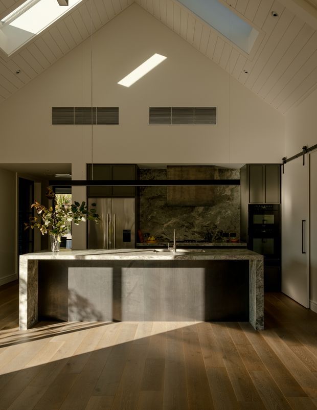 A refined kitchen viewed from the living area, featuring a large monolithic stone island with integrated sink, dark built-in cabinetry, stainless-steel refrigerator and ovens, and a dramatic stone backsplash. The room has a high pitched timber-lined ceiling with skylights and linear ventilation grilles, while warm sunlight falls across the timber flooring and island.