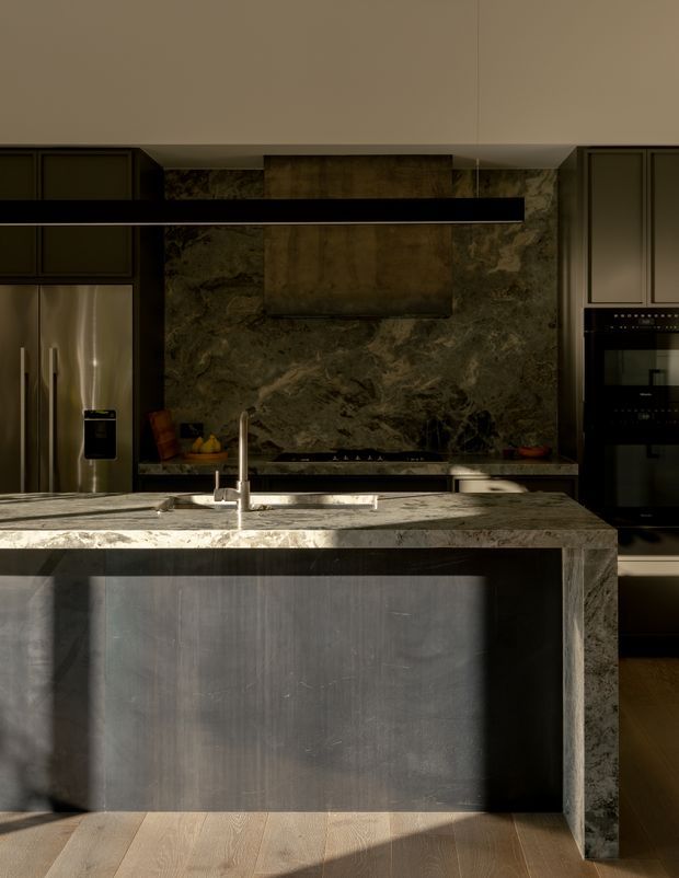 A moody, contemporary kitchen viewed directly toward a large central island. The island features a dark waterfall-edge stone benchtop, integrated sink and brushed metal tap, with dark cabinetry below. Behind it is a dramatic dark veined stone splashback and rangehood, flanked by stainless-steel refrigeration and dark built-in ovens. Warm timber flooring and strong directional sunlight create a refined, high-contrast atmosphere.