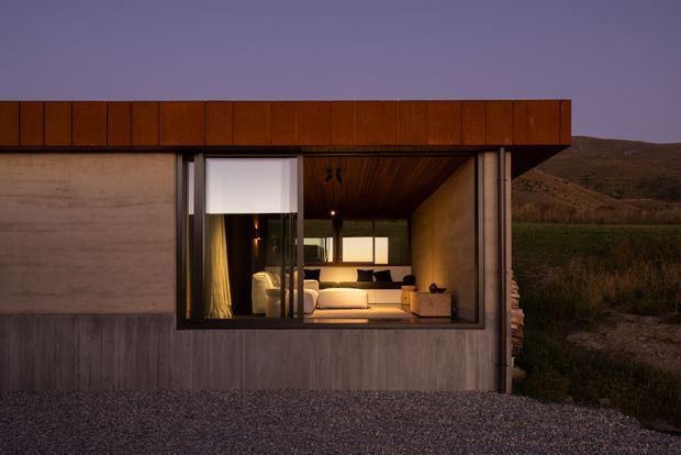 A low, modern single-storey dwelling viewed from outside at dusk, set against an open rural landscape. The facade combines a broad timber-clad flat roof edge with raw concrete walls and a large glazed opening revealing a warmly lit living room with a sofa and built-in interior finishes. A timber-lined soffit extends over the opening, and gravel surrounds the building.