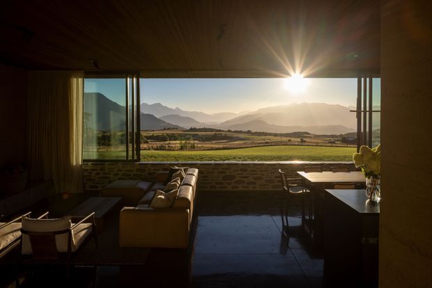 A dark, warmly finished living and dining space opens through broad floor-to-ceiling glazing to a sunlit rural valley and mountain landscape. The room features a timber-lined ceiling, exposed stone masonry below the windows, polished dark flooring, a low sofa, lounge chairs, and a dining table. Strong sunset glare creates a dramatic, contemplative atmosphere.