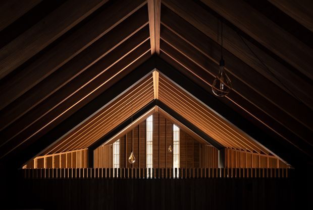 A dramatic symmetrical interior view looking toward a tall gabled end wall. Dark timber roof structure and exposed rafters frame warm-toned timber lining, with a narrow vertical glazed or translucent opening at the centre and timber balustrading in the foreground. Pendant lights hang within the vaulted space, creating a moody, refined atmosphere.