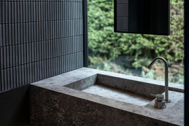 Close-up of a refined kitchen or wet-area sink set into a substantial grey natural-stone benchtop and backsplash. The basin and surround appear carved or fabricated from matching stone, paired with a slim brushed-metal gooseneck tap and minimalist soap dispenser. Dark vertically fluted wall tiles and a black-framed window overlook dense greenery, creating a moody, nature-connected interior.