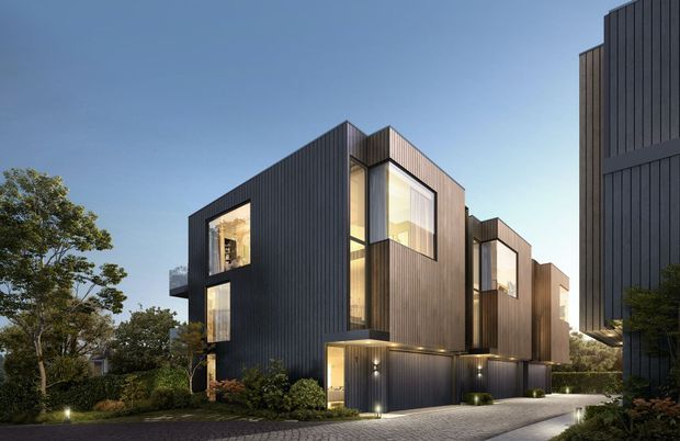 Contemporary multi-storey residential development composed of several angular volumes with dark vertical cladding, warm timber-toned facade sections, and large illuminated windows. The buildings are arranged along a landscaped paved driveway with integrated garage or entry-level openings, garden planting, and subtle exterior lighting, shown at dusk.