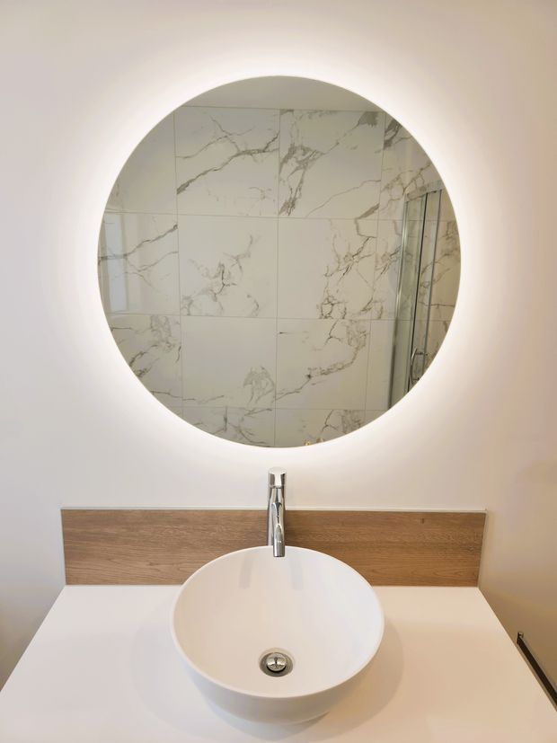 A contemporary bathroom vanity featuring a round backlit mirror, white vessel basin, chrome tapware, white benchtop, and a horizontal timber-look backsplash panel. The mirror reflects a shower area clad in large-format white marble-look tiles with grey veining.