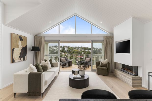 Bright contemporary living room with a vaulted white timber-lined ceiling and prominent triangular clerestory glazing. The room has pale timber flooring, white walls, a large cream sofa, dark accent armchairs, a round black coffee table, a wall-mounted television, and a linear fireplace within built-in joinery. Sliding glass doors open to a balcony overlooking a leafy residential landscape.