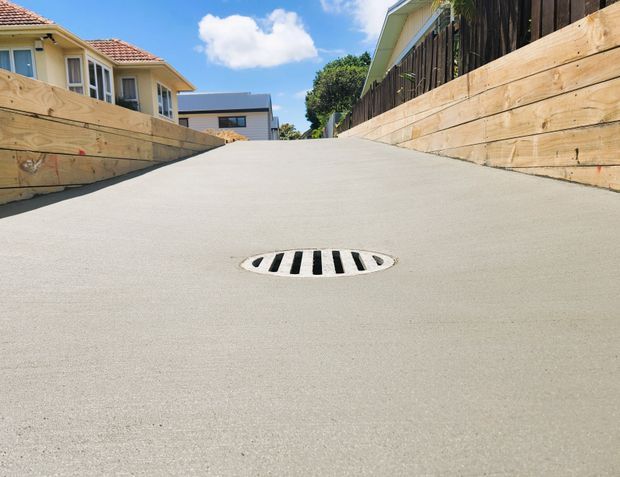 A low-angle view up a newly finished concrete driveway or access ramp, bordered on both sides by timber retaining walls and fencing. Residential houses are visible at the top of the incline beneath a bright blue sky. A central oval stormwater drain grate is set into the smooth concrete surface.