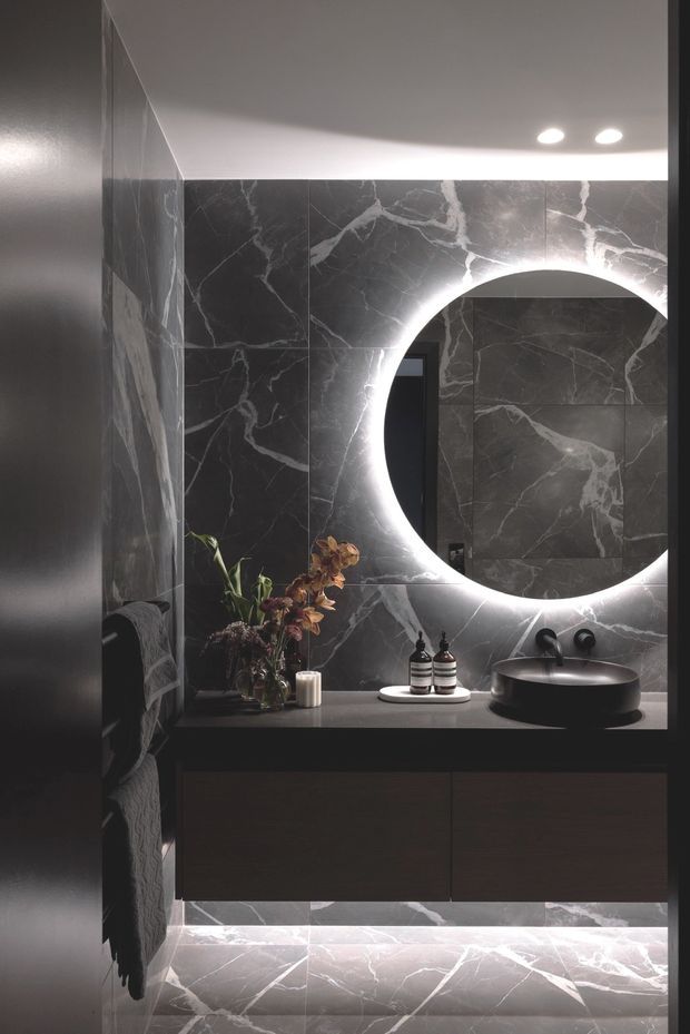 A dark, luxurious bathroom vanity with large-format charcoal marble-look tiles featuring prominent white veining. The space includes a floating dark vanity, dark round vessel basin, wall-mounted black tapware, a large backlit circular mirror, under-vanity lighting, recessed ceiling downlights, and decorative accessories.