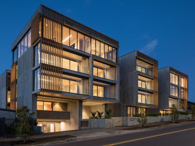 Twilight exterior photograph of a contemporary multi-storey residential development, with several stepped cubic buildings, extensive floor-to-ceiling glazing, concrete walls, and vertical timber screening. Warm interior lighting contrasts with the deep blue sky, while landscaped entry areas and a street frontage sit in the foreground.