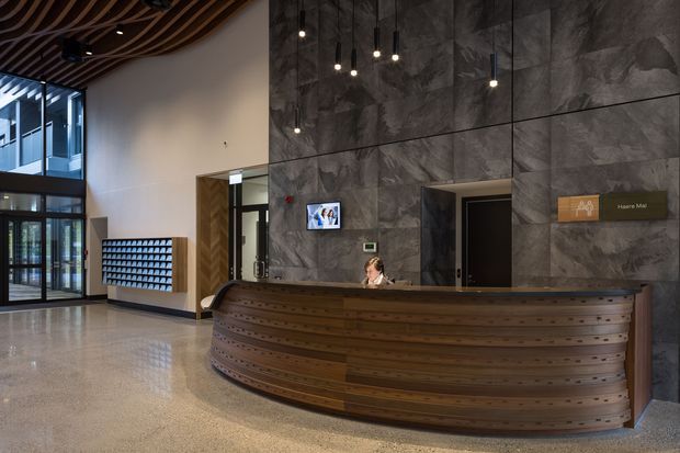 A spacious contemporary reception lobby dominated by a long, curved timber reception desk. The desk sits against a dramatic dark textured stone feature wall with suspended pendant lights and a small reception workstation. The lobby has polished terrazzo or concrete flooring, extensive glazed entry doors, a sculptural curved timber slatted ceiling, and a wall-mounted directory sign. The overall design is refined, modern, and hospitality-oriented.