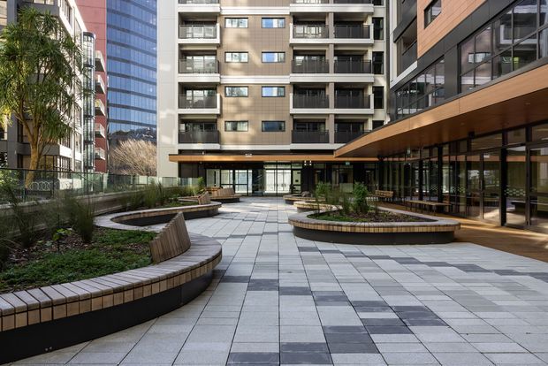 A landscaped urban courtyard framed by contemporary multi-storey apartment and commercial buildings. The paved plaza uses large light-grey and dark-grey rectangular tiles, with curved built-in timber-toned bench planters, low greenery, and mature palms. Ground-floor glazed frontage and projecting upper-level balconies surround the open communal space.