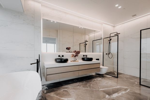 A bright, contemporary bathroom featuring a double vanity with floating wood-look drawers, two black vessel basins, wall-mounted black fixtures, large mirrors, a freestanding white bathtub, and a frameless glass shower enclosure with black metal framing. Large marble-look wall and floor tiles create a refined, cohesive finish.
