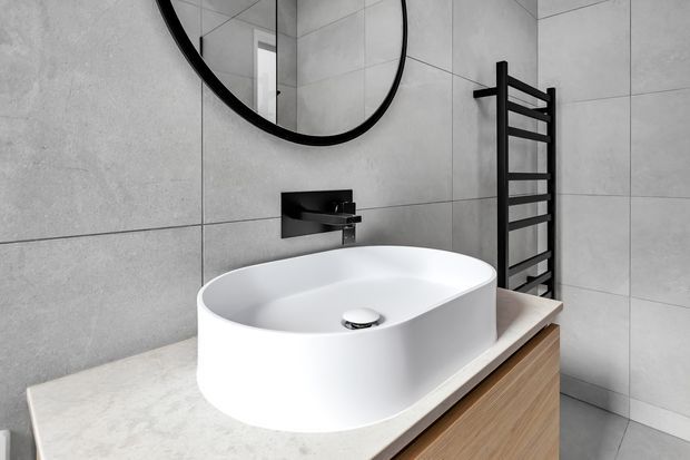 A contemporary bathroom featuring a white oval vessel basin on a pale stone benchtop with timber cabinetry below. Large light-grey tiled walls, a black wall-mounted tap, round black-framed mirror, and black ladder-style heated towel rail create a clean, monochromatic look with warm timber contrast.