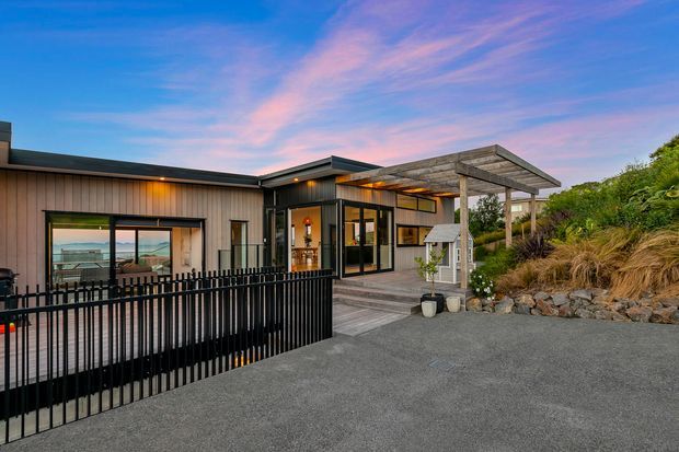 Contemporary single-storey residence viewed from the driveway, featuring dark vertical timber-style cladding, extensive glazing, a low-pitched roof, and a timber pergola over a raised deck. A black metal balustrade encloses the deck, while landscaped rockwork and grasses border the approach. The interior and deck look toward a coastal or water view visible through the glazing.