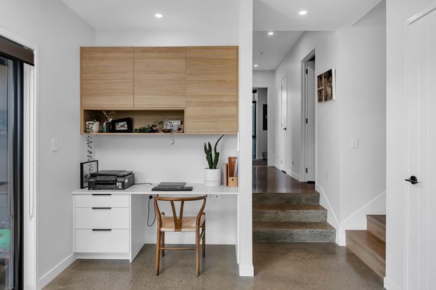 Bright contemporary home office nook integrated into a hallway, featuring a built-in white desk with drawers, overhead timber cabinetry, a wooden chair, printer, and small plants. Polished concrete flooring continues through the circulation space, with a short stairway and white doors visible beyond.