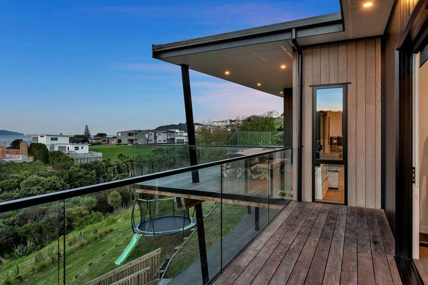 A contemporary elevated timber deck and balcony with clear glass balustrades, overlooking a landscaped suburban setting. The covered outdoor area features recessed ceiling lights, timber wall cladding, dark structural posts, and a sliding glass door opening into the home. A trampoline and garden are visible below.
