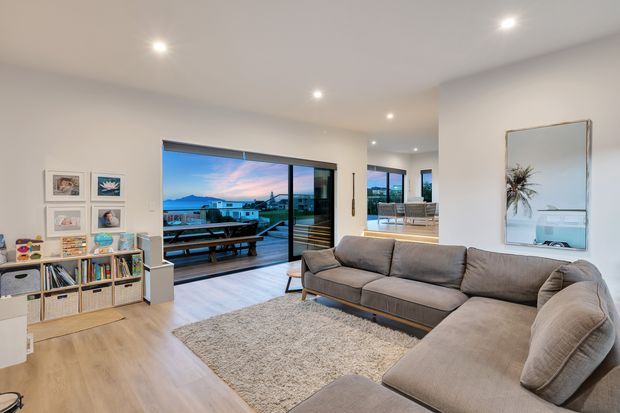 Bright contemporary living room with a large L-shaped grey sectional sofa, textured rug, light timber flooring, and a low open shelving unit filled with books and decor. Floor-to-ceiling sliding glass doors open to a deck and provide expansive views, while recessed ceiling lights and neutral finishes create an airy, relaxed interior.