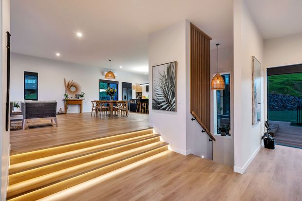 Bright contemporary open-plan interior with warm timber flooring, a raised dining and living area reached by illuminated stepped flooring, and a timber slatted stair divider with black handrail. The space includes a dining table, pendant lighting, artwork, recessed downlights, and extensive glazing opening toward an outdoor deck.