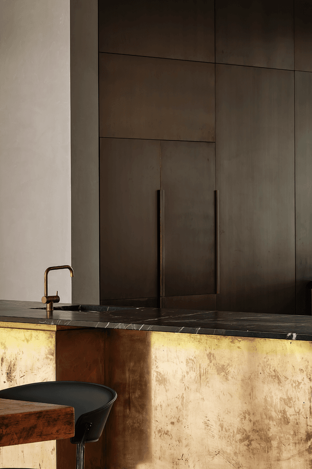 A moody, close-up view of a sophisticated kitchen or bar area. Tall dark bronze-brown flat-panel cabinetry with slim vertical handles forms the backdrop, while a dark stone benchtop incorporates an undermount sink and a brushed brass gooseneck tap. The island or counter front is clad in distressed, reflective brass-toned metal, with a dark sculptural bar stool and timber tabletop in the foreground. The palette is warm, dark, and luxurious with an industrial character.