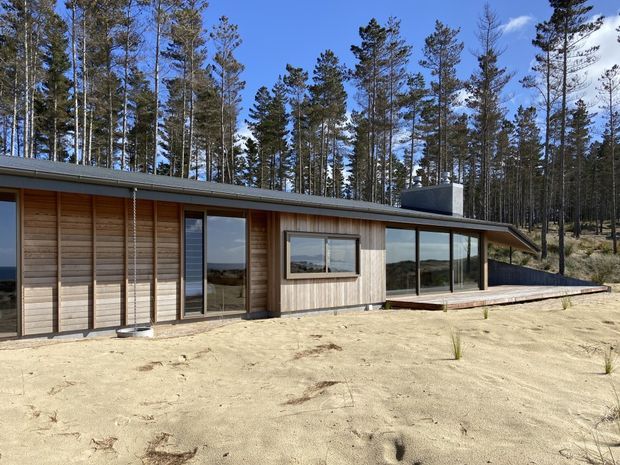 Single-storey low-profile house set in a pine-forested, sandy landscape. The exterior has extensive horizontal timber cladding, dark-framed glazing, a shallow sloping roof, and a long timber deck along the glazed living area. The simple linear form and strong connection to the surrounding woodland are prominent.