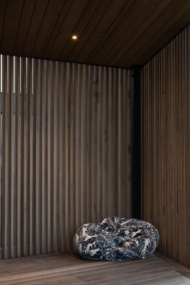 A dark, intimate interior corner clad almost entirely in vertically ribbed timber wall panels and matching timber ceiling boards. The timber flooring continues across the space, with a single recessed downlight overhead and a large low, rounded beanbag-style lounge seat upholstered in a dark botanical pattern. The overall composition is textural, warm, and minimalist, with strong vertical rhythm and subdued lighting.