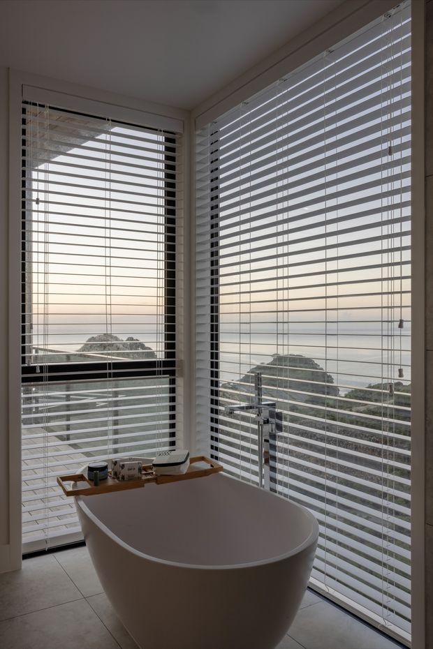 A modern bathroom with a freestanding oval bathtub positioned beside large corner windows. White horizontal venetian blinds cover the glazing, offering views toward a calm coastal or waterfront setting. Pale tiled flooring, light walls, and minimal white sanitaryware create a clean, serene atmosphere. A small wooden bath tray rests across the tub, and a chrome floor-mounted tap is visible beside it.