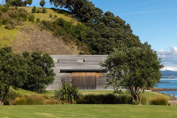 A low-profile contemporary building nestled into a landscaped coastal hillside. The facade is clad in horizontal weathered timber boards, with a large dark timber entrance door and subtle roof-level skylight elements. Native planting, lawn, a bay or ocean inlet, and distant hills are visible around the structure.