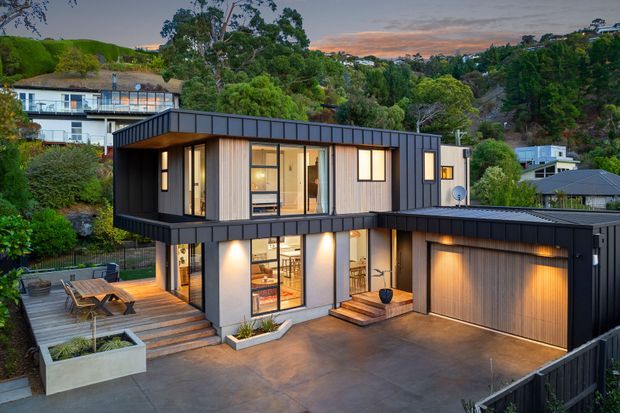 A contemporary two-storey residence on a landscaped suburban hillside, featuring dark standing-seam metal cladding, light vertical timber-look wall panels, extensive glazing, projecting upper-level forms, and an attached double garage. A timber deck and outdoor dining area sit beside the home, with warm interior and exterior lighting visible at dusk.