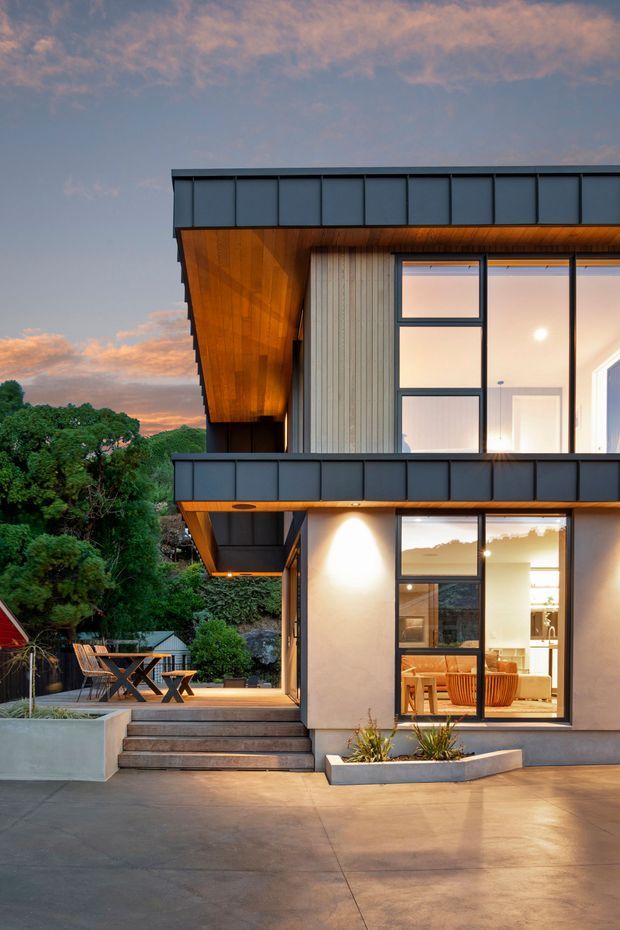Contemporary two-storey house exterior at dusk, featuring dark metal cladding and parapet edges, warm timber soffits, vertical timber wall cladding, and expansive black-framed glazing. A covered outdoor dining terrace and stepped concrete-paved forecourt are visible, with warm interior lighting glowing through the windows.