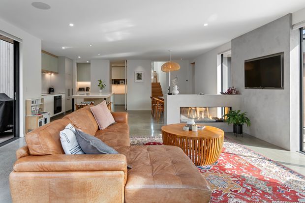 A bright, open-plan contemporary living room with a large tan leather sectional sofa and chaise, patterned red Persian-style rug, round wooden coffee table, and a concrete-look feature wall incorporating a fireplace and television. The room opens directly into a dining area and streamlined white kitchen, with timber stairs and large glazed doors adding depth and natural light.