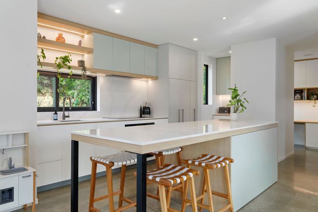 Bright contemporary kitchen with a large white island featuring an integrated breakfast bar and four timber stools. Pale flat-panel cabinetry runs along the rear wall, with open display shelving, a black-framed window, white benchtops, and subtle timber accents. The space is clean, airy, and softly lit, with a pantry or utility area visible beyond the kitchen.
