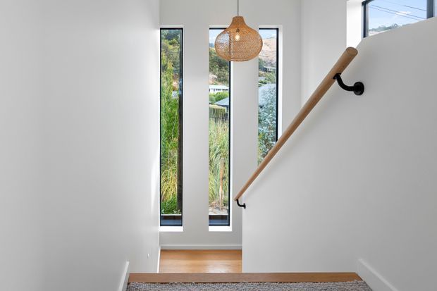Bright minimalist stairway with white walls, narrow vertical windows, a timber handrail on black brackets, pale timber flooring, and a woven pendant light. A landscaped suburban exterior is visible through the glazing.