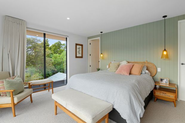 A bright, calm bedroom with a large glazed window overlooking greenery, pale carpet, a neatly made bed with a woven rattan headboard, and a muted sage-green vertical-panel feature wall. Light timber and upholstered furniture, soft neutral bedding, blush and tan cushions, sheer curtains, and warm pendant lights create a relaxed contemporary interior.