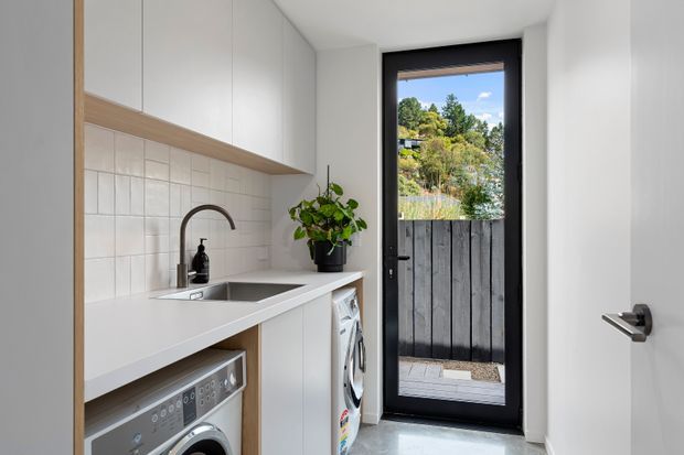 Bright, compact contemporary laundry room with white built-in cabinetry, a pale benchtop, tiled splashback, stainless sink and black tapware. A front-loading washing machine and dryer are integrated beneath the counter, with a black-framed glazed door opening to a small fenced outdoor area.