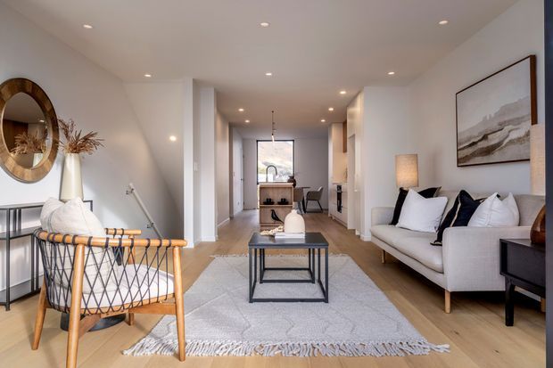Bright, narrow contemporary open-plan living room with a timber floor, white walls and recessed ceiling lighting. The room includes a white sofa with dark and white cushions, a black-framed coffee table, a textured light rug, a woven timber-framed armchair, decorative wall mirror, artwork and a small dining or kitchen area visible at the rear.