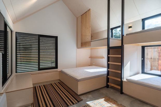 Compact, highly functional bedroom with built-in bunk beds and a raised double bed, pale timber cabinetry, open shelving, black-framed windows, integrated horizontal blinds, and a central black ladder and storage unit. The room has a warm neutral palette, streamlined built-in joinery, and a striped area rug.
