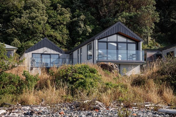 Exterior view of a contemporary gabled house set into a densely vegetated, sloping coastal site. The building has dark vertical cladding, large floor-to-ceiling glazed façades, projecting angular roof forms, and a raised lower level. Dry grasses and rocky shoreline vegetation occupy the foreground.