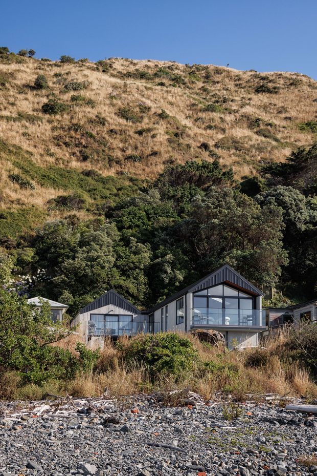 A modern group of small gabled buildings set into a densely vegetated hillside above a rocky beach. The main structure has dark vertical cladding, a pitched gable roof, and a large glazed facade with a projecting upper-level balcony or living area. The surrounding landscape includes native-looking shrubs, dry grass, steep terrain, and shoreline stones beneath a clear blue sky.