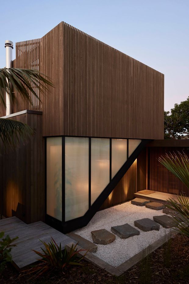 Exterior view of a contemporary multi-level building with a strongly articulated vertical timber screen facade. Black-framed translucent glazing forms an angled lower-level volume, while a gravel courtyard with large stepping stones, timber decking, and tropical planting surrounds the structure. Warm interior lighting is visible through the glazing, and a metal flue projects above the roofline.