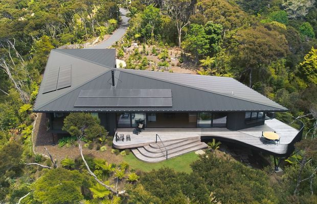 Aerial view of a contemporary single-storey house nestled in dense native bushland. The home has a broad dark grey metal roof with multiple sloping planes, dark exterior walls, extensive glazing, and a large light-toned timber or composite deck wrapping around the front and side. Curved steps lead from the deck to a landscaped garden, with a winding driveway or walkway visible through the trees.