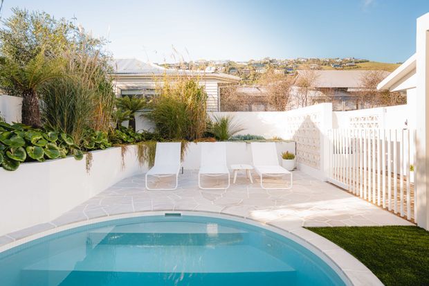 Bright outdoor courtyard terrace with a curved swimming pool, pale stone paving, white sun loungers, white privacy fencing, and lush layered planting in raised garden beds. The scene overlooks a suburban hillside and features a clean, contemporary coastal feel.