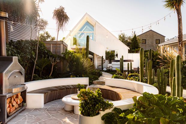 A sunny landscaped courtyard and outdoor entertaining area in front of a distinctive white gabled house. The space features curved built-in seating around a circular fire pit or table, pale irregular stone paving, abundant palms, succulents and tall cacti, festoon lighting, and a built-in outdoor fireplace at left. The house has a sharply pitched gable with large glazing and white cladding, creating a relaxed modern resort-like setting.