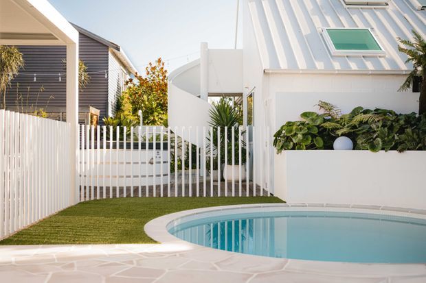 Bright architectural exterior view of a compact residential courtyard centred on a curved swimming pool. White rendered and vertically ribbed walls, a sculptural curved stair or ramp enclosure, white vertical-bar fencing, pale paving, landscaped garden beds, and a neighbouring dark weatherboard structure create a clean contemporary composition.