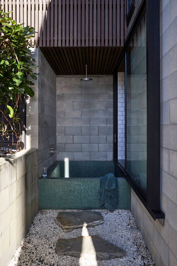 A narrow, enclosed outdoor bathing courtyard with a tiled plunge pool or bath at the far end, surrounded by grey concrete block walls. Dark timber slats form a sheltered overhead screen, while black-framed glazing runs along the right side. Green mosaic tiles line the pool and raised ledge, with white gravel and irregular stepping stones in the foreground. A wall-mounted shower and small basin tap are visible, creating a private, minimalist Japanese-inspired garden bathing space.