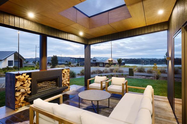 A sophisticated covered outdoor lounge with floor-to-ceiling glazing, warm timber-lined ceiling and walls, a central skylight, and a built-in fireplace with stacked firewood. White upholstered seating surrounds a round coffee table on a timber deck, with views across a landscaped yard and lake beyond.