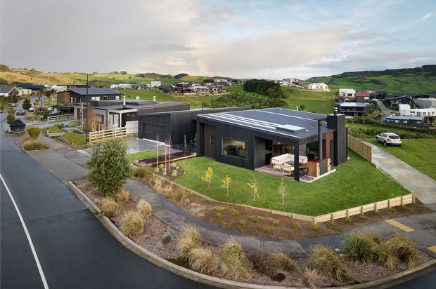 Aerial view of a contemporary single-storey residential home in a suburban development. The dark charcoal-clad house has a low-pitched metal roof, extensive glazing, a paved driveway, landscaped front garden, timber fencing and an outdoor entertaining area. Rolling green hills and neighbouring homes form the background.
