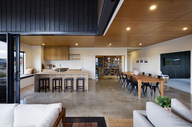 Open-plan contemporary living space combining a kitchen with island seating, dining area, and lounge. The room features polished concrete flooring, warm timber ceiling lining, a dark textured feature wall and ceiling section, light cabinetry, extensive glazing, and a built-in wine display visible beyond the dining table.