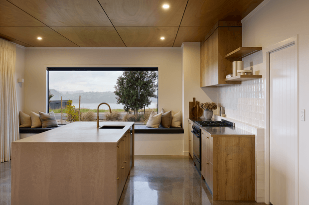 A warm, compact contemporary kitchen with a large central island, integrated sink and curved brass-toned tap, timber cabinetry, open shelving, and a white glazed tile splashback. A built-in window seat with cushions overlooks a landscaped waterfront or lakeside setting. The room has a timber-lined ceiling, polished grey concrete flooring, recessed lighting, and a built-in oven and cooktop.