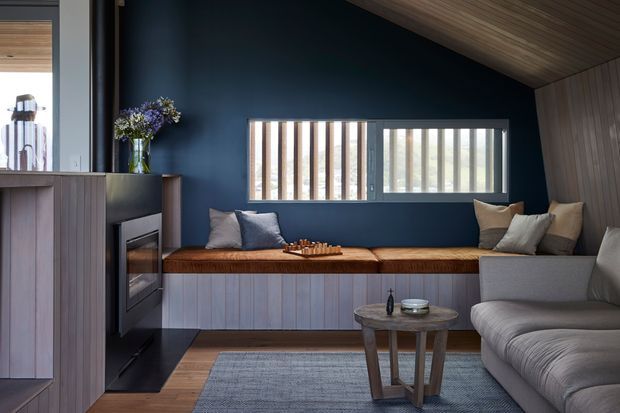 A refined contemporary living room with a built-in window seat and fireplace wall. The room features deep navy painted walls, vertical timber-clad cabinetry and bench joinery, a timber slatted ceiling, narrow clerestory-style windows with vertical privacy fins, a grey sectional sofa, and a textured grey rug. Warm timber and muted upholstery create a calm, cozy atmosphere.
